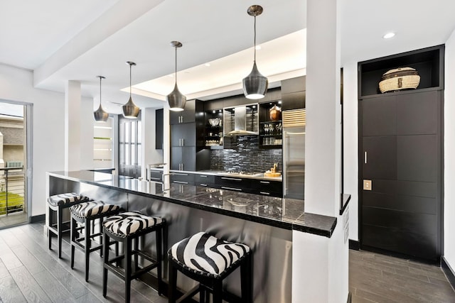 kitchen with pendant lighting, wall chimney exhaust hood, dark wood-type flooring, a kitchen bar, and decorative backsplash