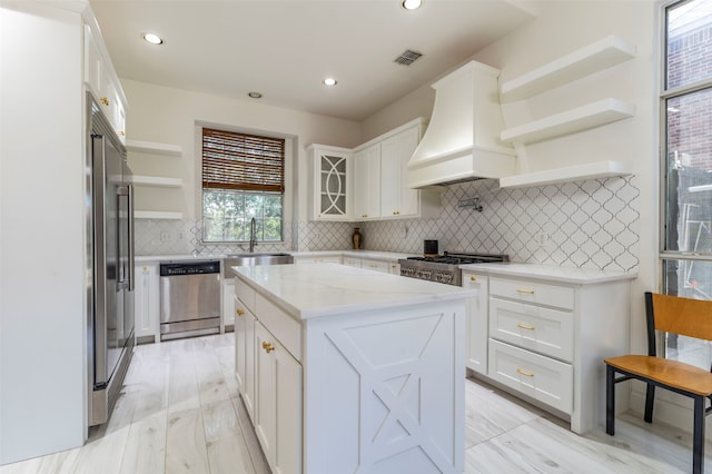 kitchen with custom range hood, appliances with stainless steel finishes, white cabinets, a center island, and sink