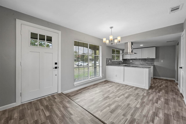 kitchen with wall chimney range hood, a notable chandelier, kitchen peninsula, white cabinets, and light wood-type flooring