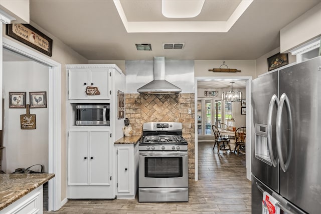 kitchen with white cabinets, pendant lighting, wall chimney range hood, stainless steel appliances, and light hardwood / wood-style floors