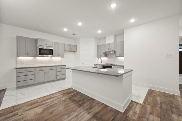 kitchen featuring built in microwave, sink, dark stone counters, gray cabinets, and a kitchen island with sink