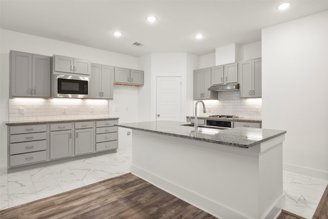 kitchen featuring built in microwave, sink, dark stone counters, gray cabinets, and a center island with sink