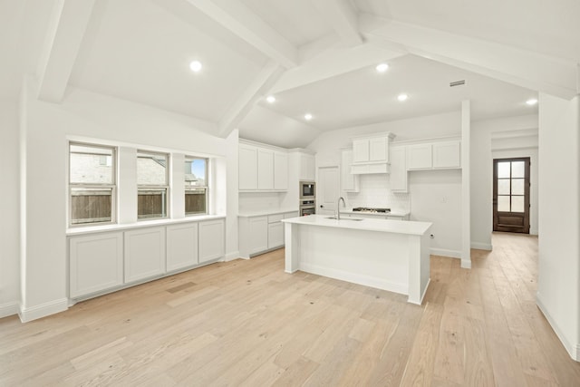 kitchen with sink, white cabinetry, vaulted ceiling with beams, light hardwood / wood-style floors, and an island with sink