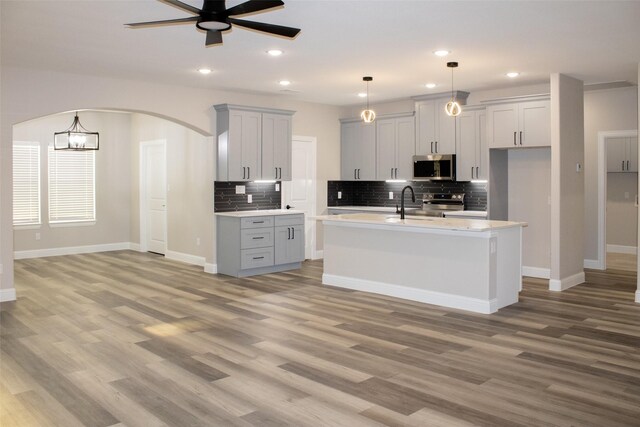 kitchen with an island with sink, decorative light fixtures, appliances with stainless steel finishes, ceiling fan with notable chandelier, and light hardwood / wood-style floors