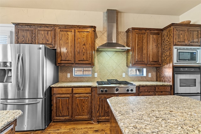 kitchen with appliances with stainless steel finishes, light stone counters, tasteful backsplash, wood-type flooring, and wall chimney range hood