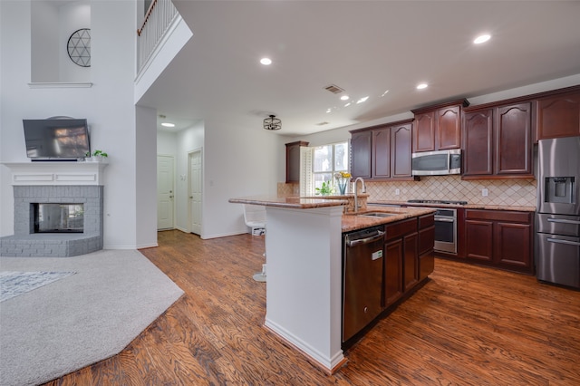 kitchen with sink, a brick fireplace, a center island with sink, dark wood-type flooring, and stainless steel appliances