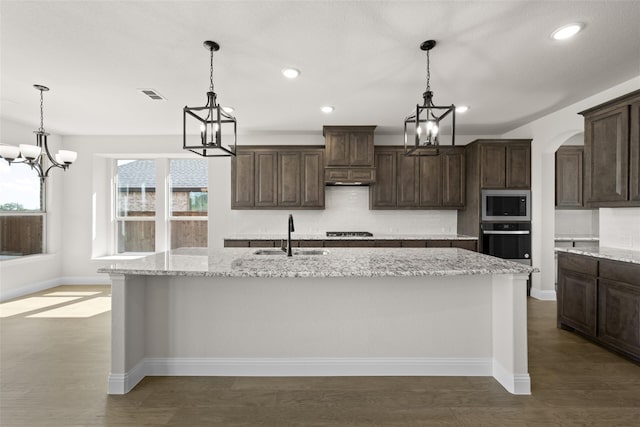 kitchen featuring sink, dark brown cabinets, a center island with sink, stainless steel appliances, and light stone countertops