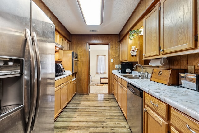 kitchen with light wood-type flooring, a textured ceiling, sink, wooden walls, and appliances with stainless steel finishes