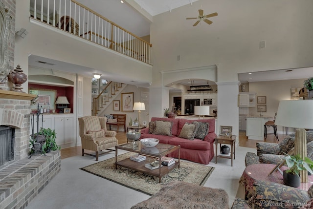 living room featuring light wood-type flooring, a brick fireplace, a towering ceiling, ceiling fan, and ornate columns