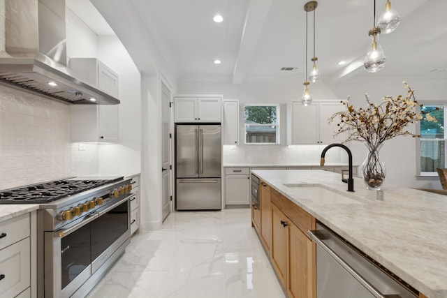 kitchen with white cabinetry, high quality appliances, wall chimney exhaust hood, light stone countertops, and decorative light fixtures