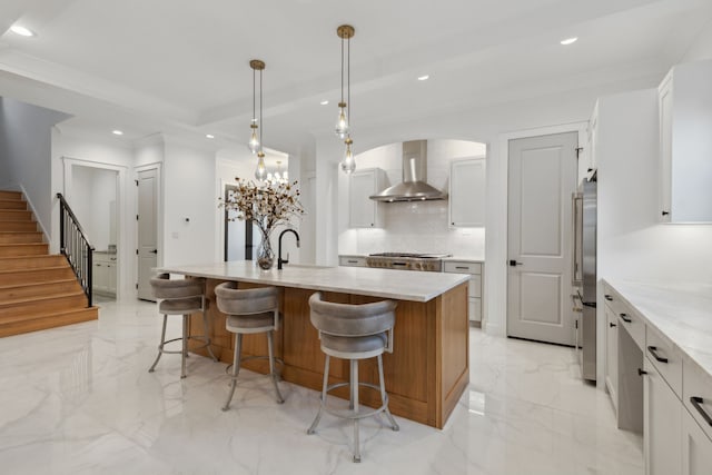 kitchen with light stone counters, an island with sink, white cabinets, pendant lighting, and wall chimney range hood