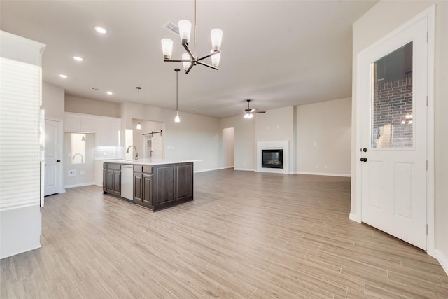 kitchen featuring stainless steel dishwasher, light wood-type flooring, an island with sink, decorative light fixtures, and dark brown cabinetry