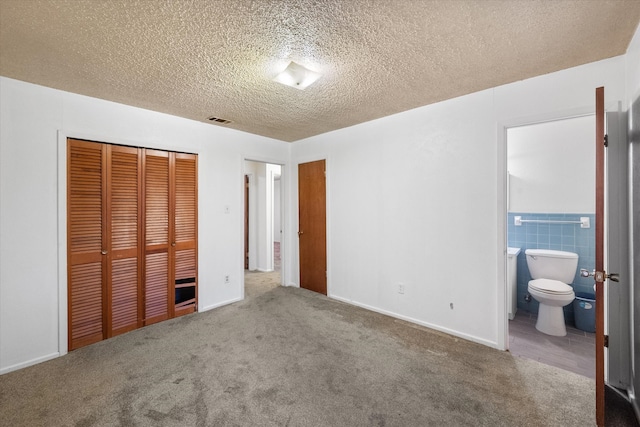 unfurnished bedroom featuring tile walls, a closet, a textured ceiling, carpet flooring, and ensuite bathroom