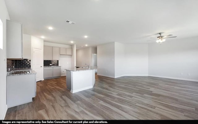 kitchen with decorative backsplash, an island with sink, dark hardwood / wood-style flooring, light stone countertops, and ceiling fan