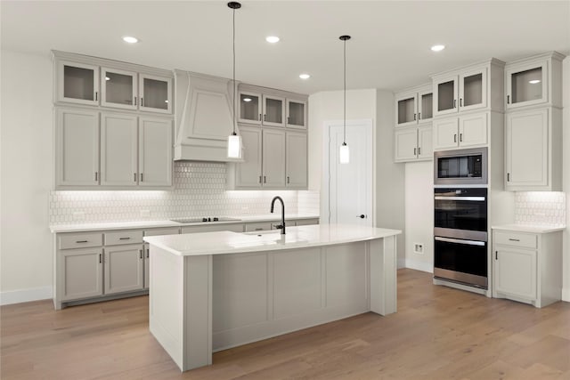 kitchen with an island with sink, appliances with stainless steel finishes, glass insert cabinets, hanging light fixtures, and light countertops