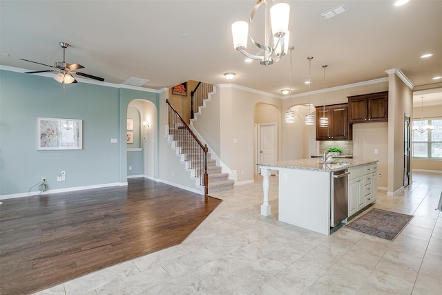 kitchen with light stone countertops, a center island with sink, ceiling fan with notable chandelier, and ornamental molding