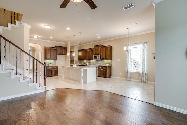 kitchen with light wood-type flooring, ceiling fan with notable chandelier, an island with sink, and hanging light fixtures