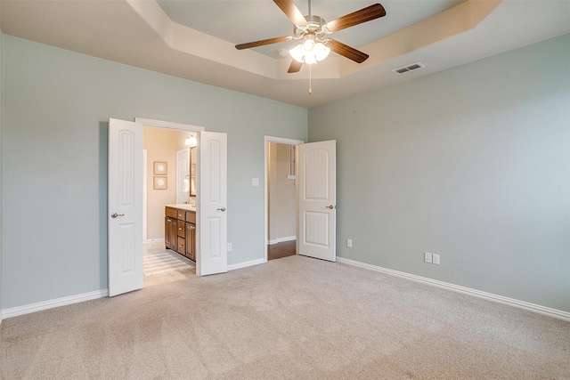 unfurnished bedroom featuring light carpet, ceiling fan, a raised ceiling, and ensuite bathroom