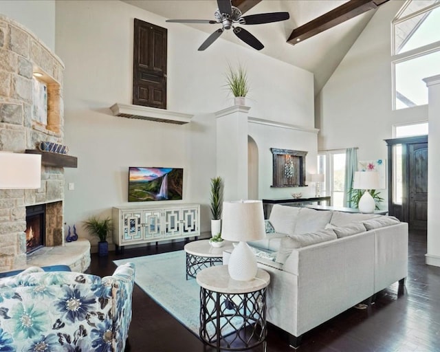 living room featuring dark hardwood / wood-style flooring, beam ceiling, high vaulted ceiling, a stone fireplace, and ceiling fan