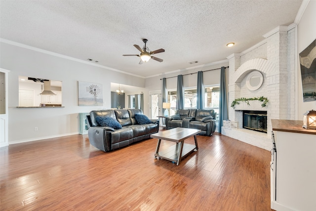 living room with ceiling fan with notable chandelier, a fireplace, a textured ceiling, wood-type flooring, and crown molding