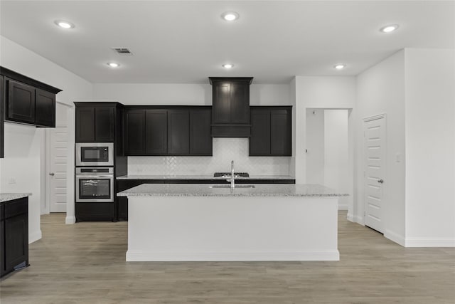 kitchen with built in microwave, light stone counters, stainless steel oven, and a center island with sink