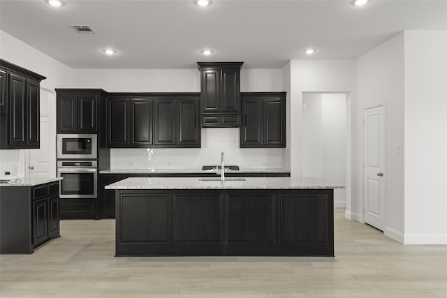 kitchen featuring backsplash, light stone counters, stainless steel appliances, a kitchen island with sink, and sink