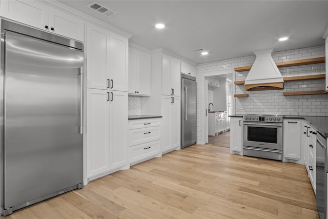 kitchen featuring appliances with stainless steel finishes, light hardwood / wood-style floors, white cabinets, backsplash, and premium range hood