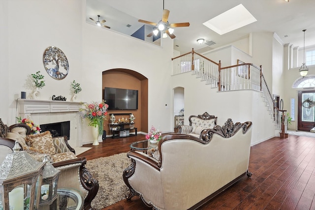 living room featuring dark wood-type flooring, a tiled fireplace, a skylight, a high ceiling, and ceiling fan