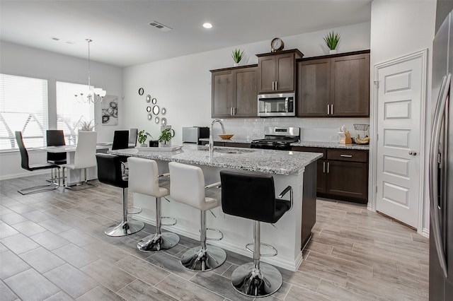 kitchen featuring sink, a kitchen island with sink, stainless steel appliances, an inviting chandelier, and light stone countertops