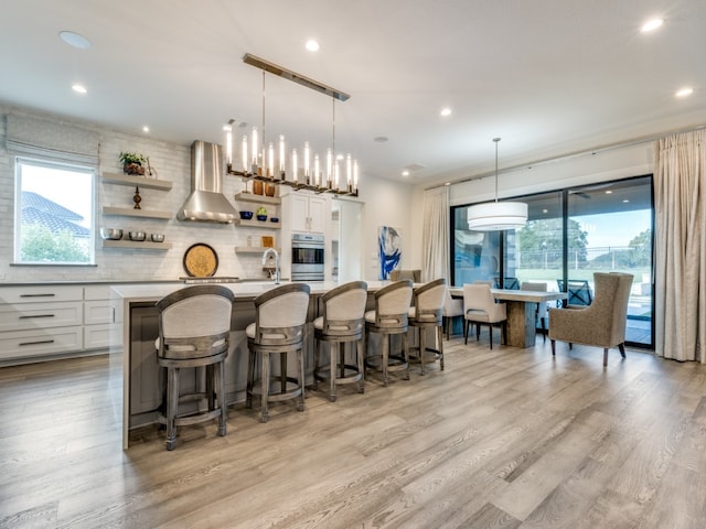 kitchen featuring hanging light fixtures, a center island with sink, white cabinetry, and wall chimney range hood