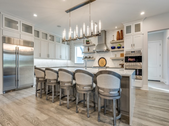 kitchen featuring appliances with stainless steel finishes, white cabinetry, wall chimney range hood, and an island with sink