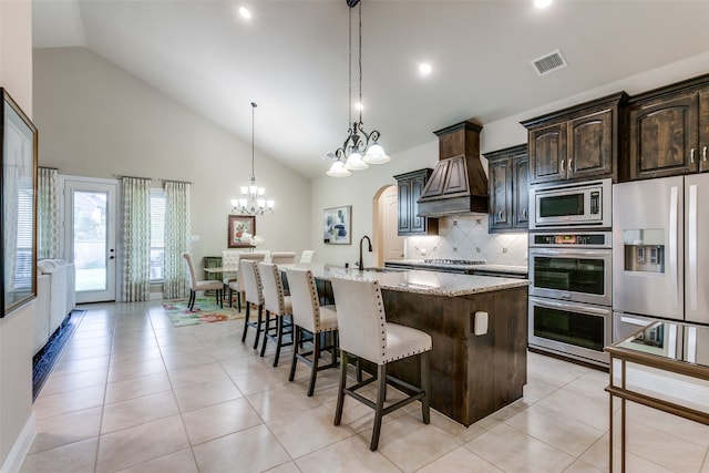 kitchen with stainless steel appliances, light stone counters, dark brown cabinetry, an island with sink, and high vaulted ceiling