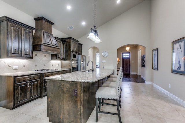 kitchen with light stone countertops, appliances with stainless steel finishes, dark brown cabinetry, and a center island with sink