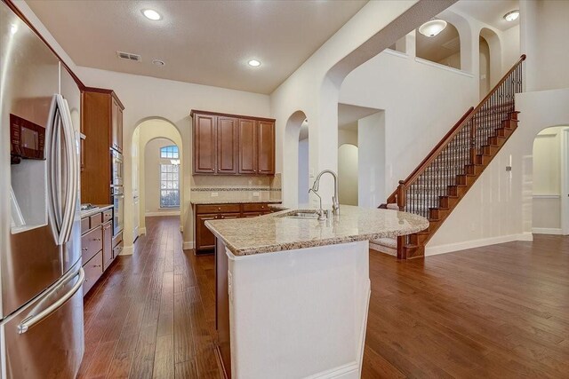 kitchen with light stone counters, an island with sink, stainless steel fridge, sink, and dark wood-type flooring