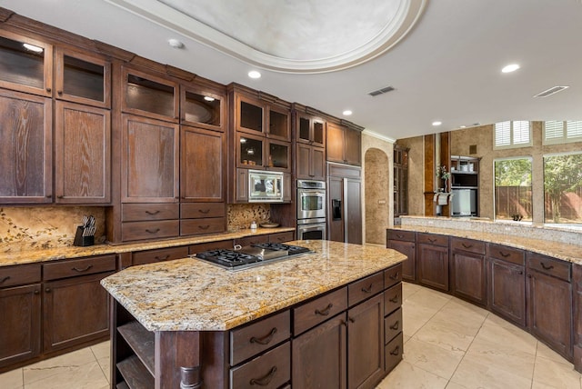 kitchen with a center island, built in appliances, decorative backsplash, light stone countertops, and dark brown cabinets