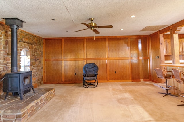 living area with a wood stove, a textured ceiling, ceiling fan, wooden walls, and light colored carpet