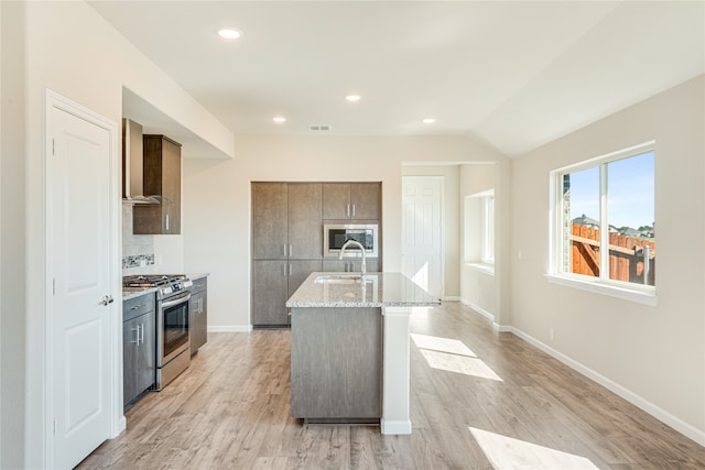 kitchen featuring wall chimney range hood, appliances with stainless steel finishes, a kitchen island with sink, light wood-type flooring, and sink