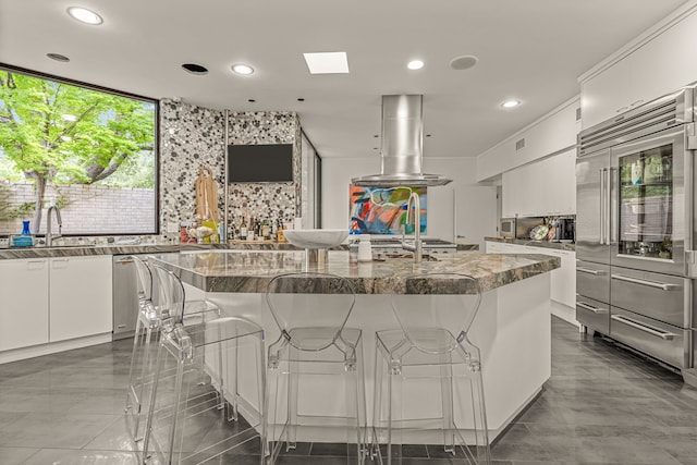 kitchen featuring white cabinets, a kitchen island with sink, island exhaust hood, a sink, and decorative backsplash