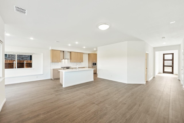 kitchen with light brown cabinets, wall chimney range hood, tasteful backsplash, light hardwood / wood-style floors, and a center island with sink