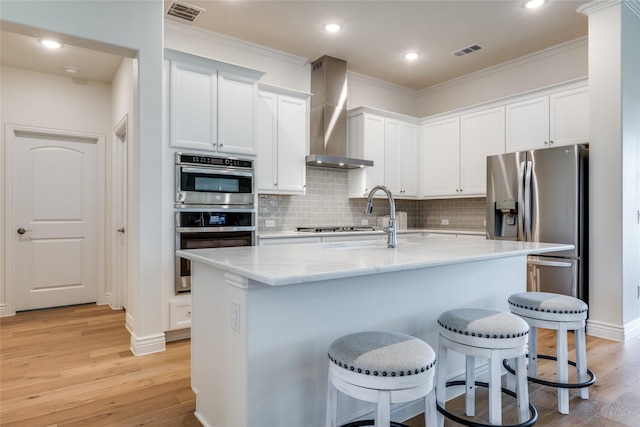 kitchen with wall chimney exhaust hood, stainless steel appliances, white cabinets, light hardwood / wood-style floors, and an island with sink