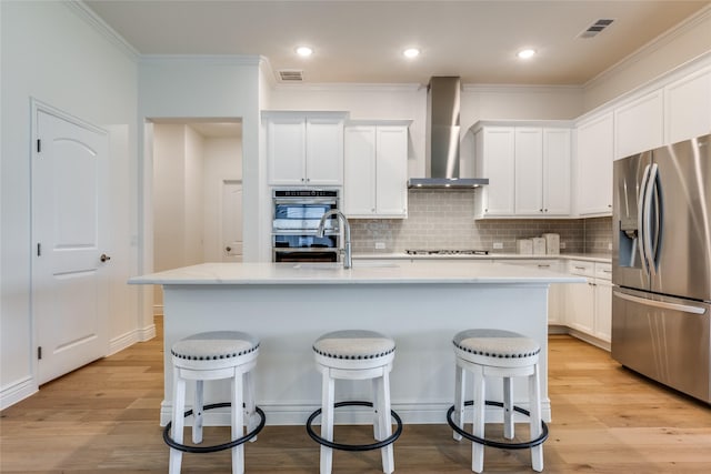 kitchen featuring appliances with stainless steel finishes, light wood-type flooring, wall chimney range hood, white cabinets, and an island with sink