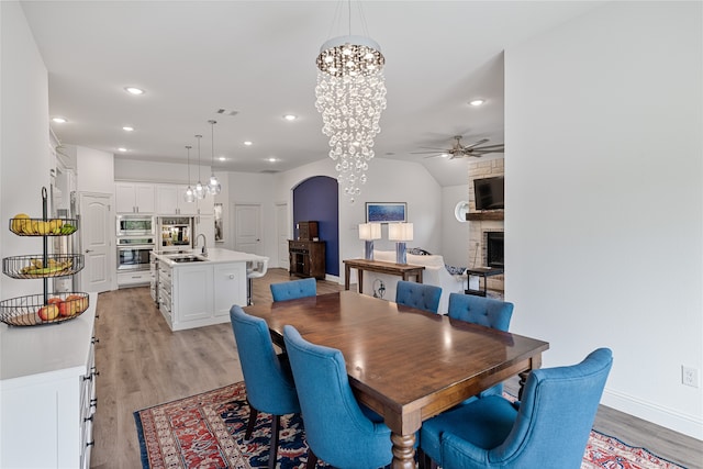 dining area featuring lofted ceiling, sink, a large fireplace, light hardwood / wood-style floors, and ceiling fan with notable chandelier