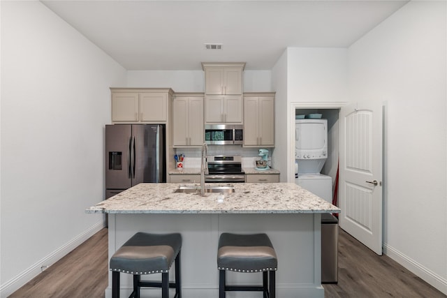 kitchen with stainless steel appliances, light stone countertops, stacked washer and dryer, and a center island with sink