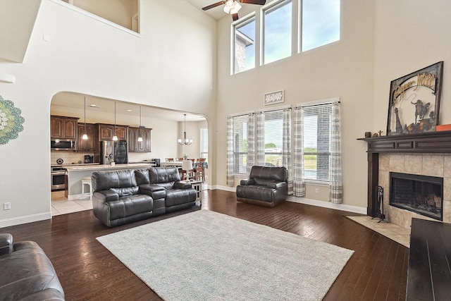 living room with ceiling fan with notable chandelier, a tile fireplace, a towering ceiling, and wood-type flooring