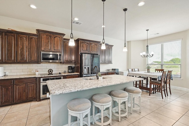 kitchen with appliances with stainless steel finishes, hanging light fixtures, an island with sink, light stone countertops, and dark brown cabinetry