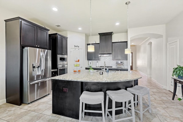 kitchen with appliances with stainless steel finishes, light stone countertops, an island with sink, backsplash, and pendant lighting