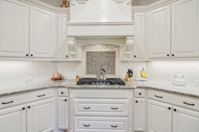 kitchen with decorative backsplash, light stone countertops, premium range hood, white cabinetry, and stainless steel gas stovetop