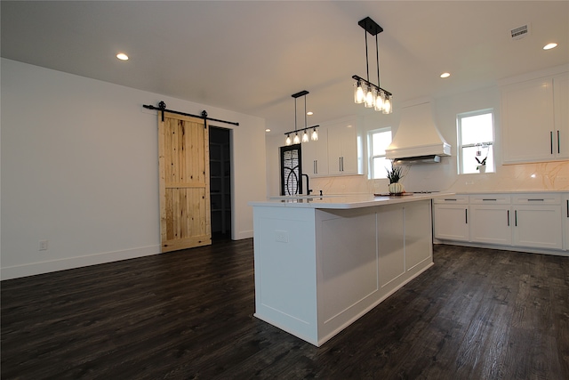 kitchen featuring an island with sink, dark wood-type flooring, white cabinetry, custom range hood, and a barn door