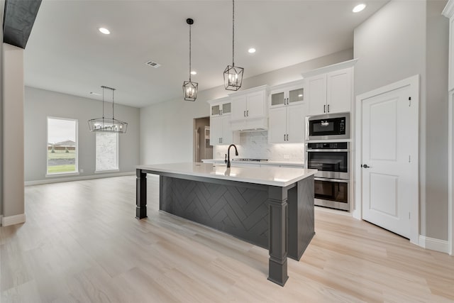 kitchen with light hardwood / wood-style floors, white cabinetry, stainless steel microwave, a center island with sink, and decorative light fixtures