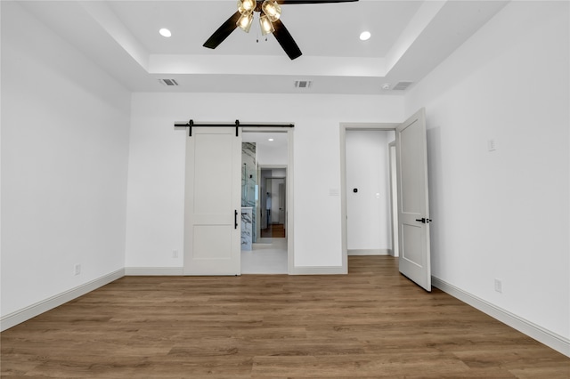 unfurnished bedroom featuring ceiling fan, a tray ceiling, hardwood / wood-style floors, and a barn door